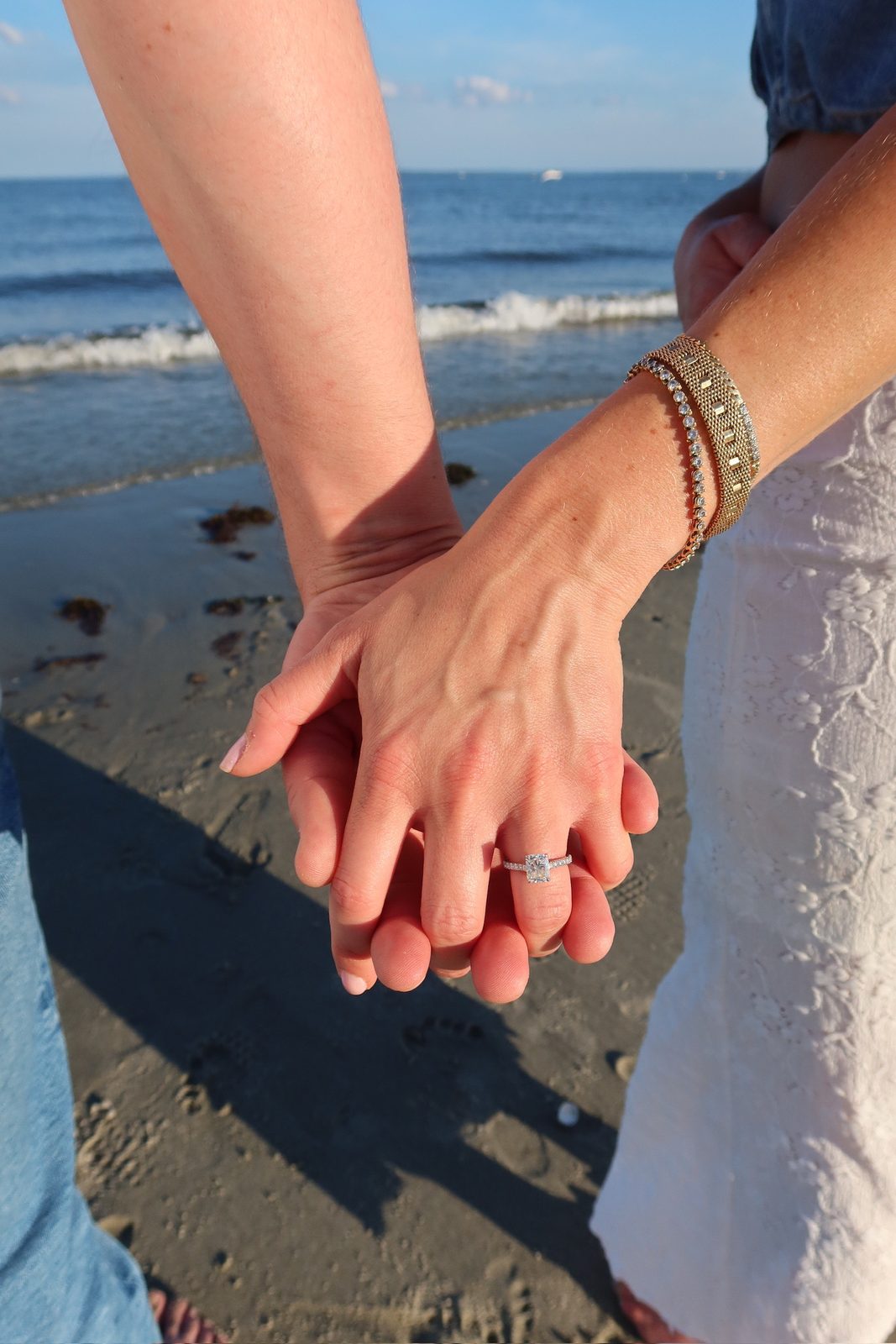 Close-up of Andrew and Jenna's clasped hands showing the engagement ring, ocean behind